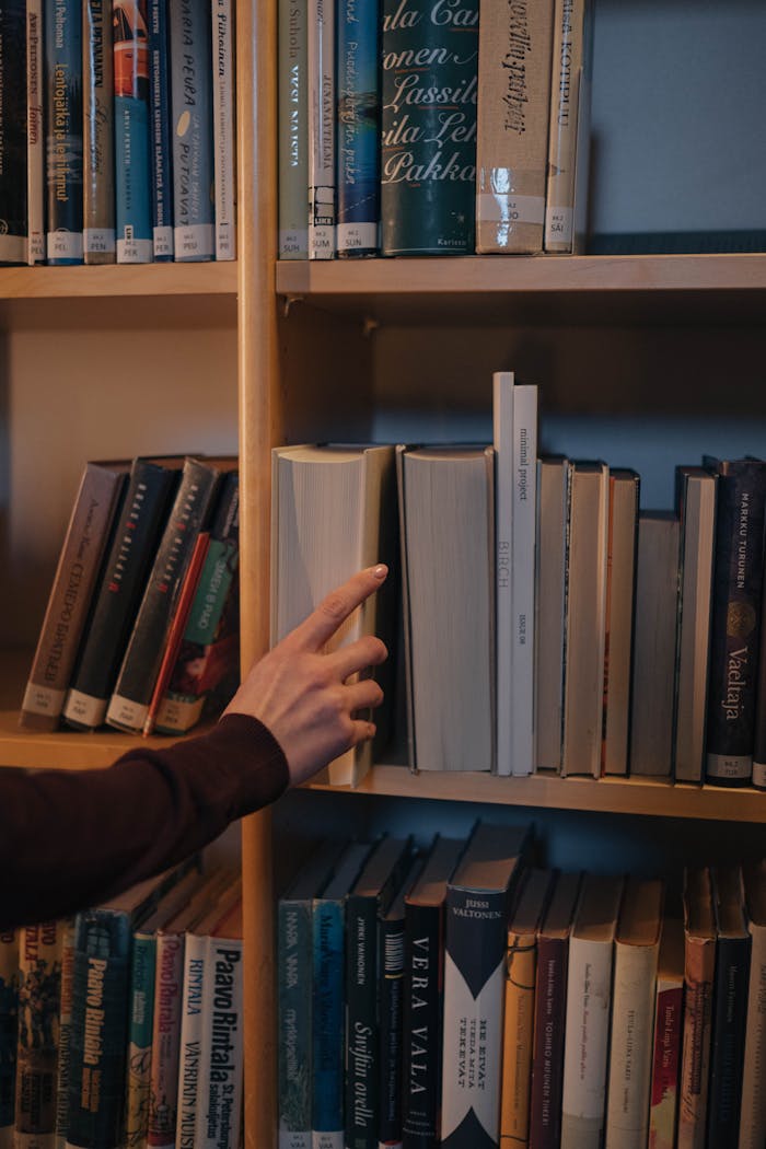 A hand reaches for a book on a neatly organized wooden bookshelf.
